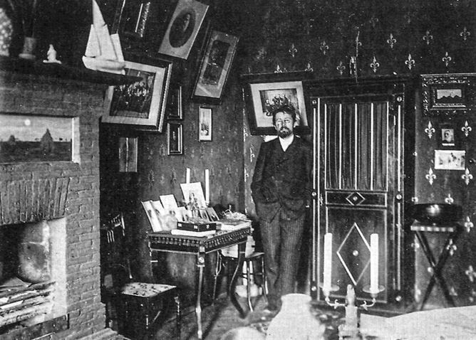 Anton Chekhov standing in his writing study at the White Dacha, surrounded by bookshelves, framed photographs on the walls, and writing materials on a desk